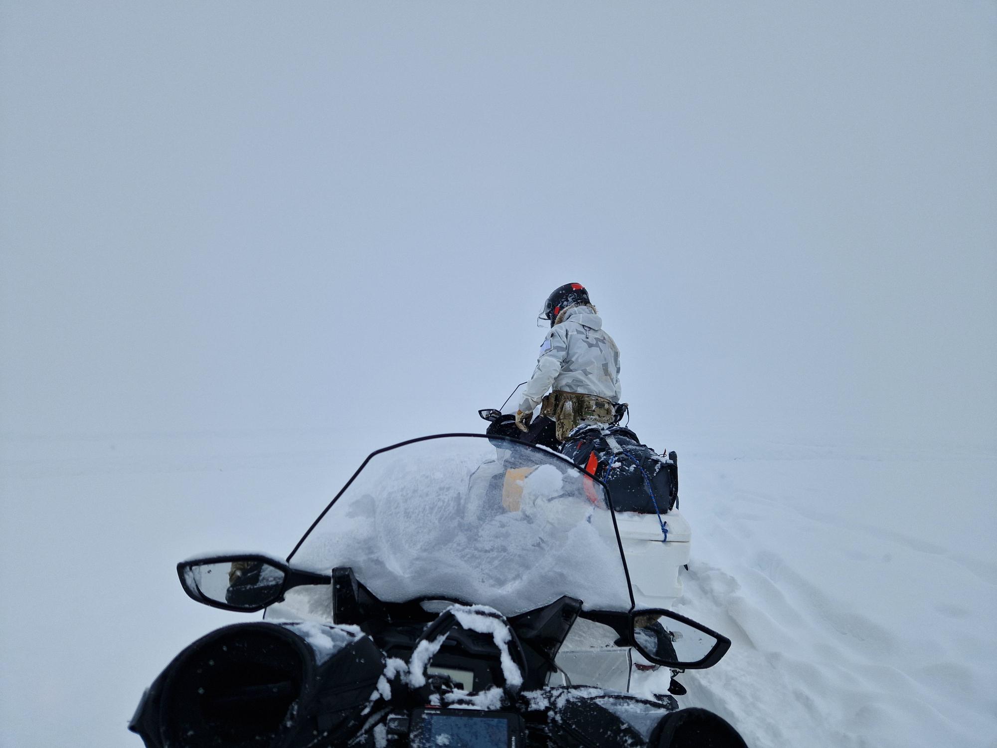 Snescooterfører i hvidt camouflagetøj sidder på snescooter i snedækket landskab med lav sigtbarhed.