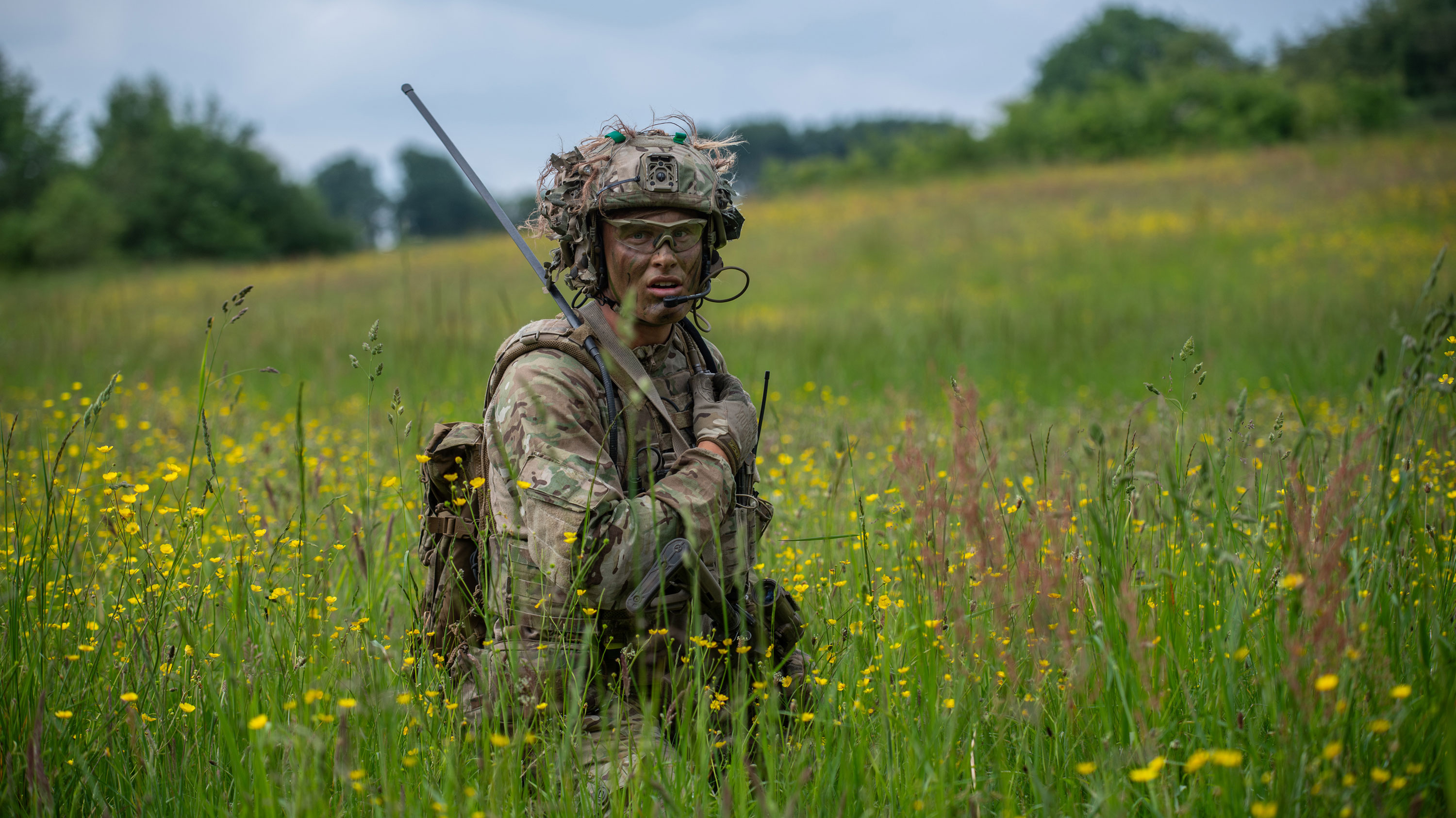 Soldat i camouflageuniform med radio og taktisk hjelm står i græs og gule vilde blomster under en overskyet himmel.