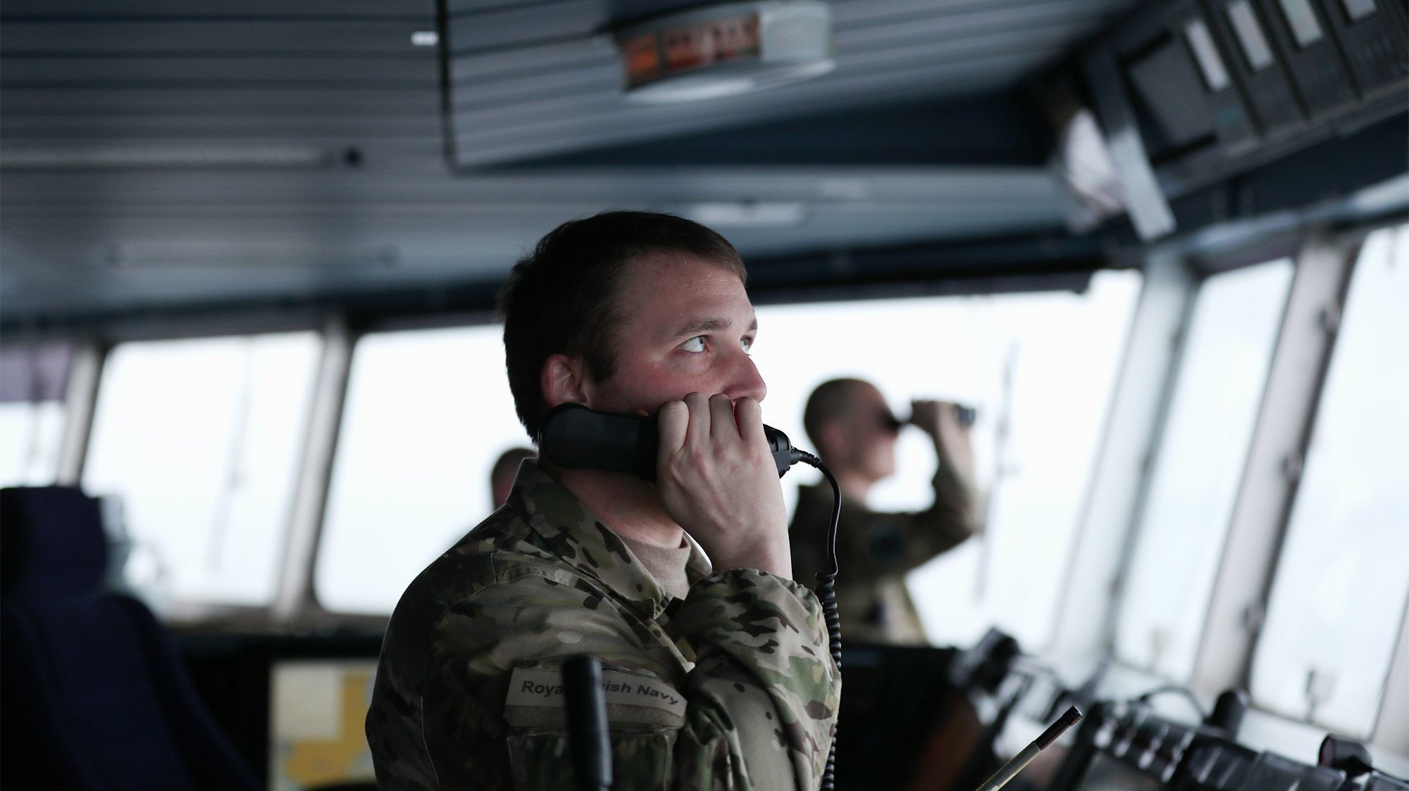 Soldat fra Søværnet taler i telefon på skibets bro, mens en kollega med kikkert holder udkig i et moderne kontrolrum.