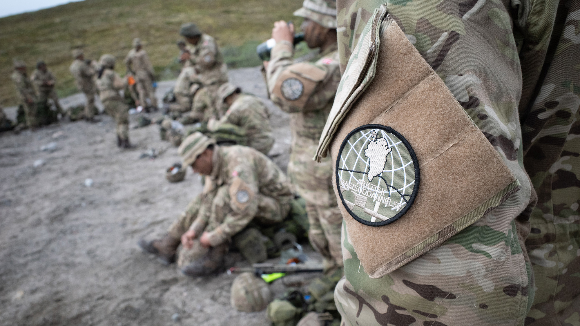 Soldater i camouflageuniform holder pause i terrænet, mens et tæt udsnit viser et skuldermærke med logoet for Arktisk Basisuddannelse.