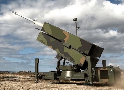 Missilsystem i camouflagefarver peger mod himlen og står opstillet på åben, sandet terræn under en skyet himmel.