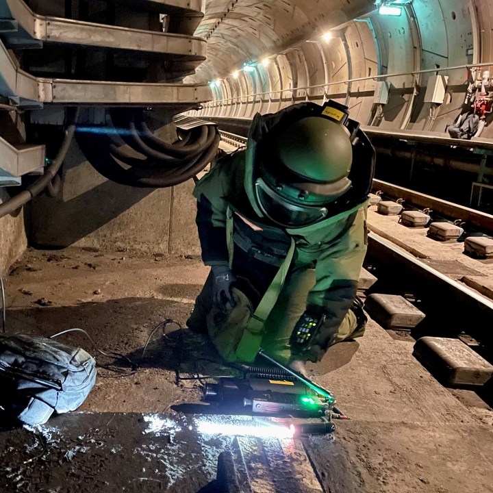 Ammunitionsrydder i beskyttelsesdragt ligger på knæ og kigger ind under Metro tog