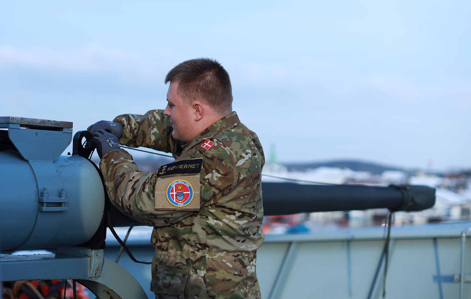 Søværnssoldat i camouflageuniform og kasket står foran moderne dansk krigsskib.