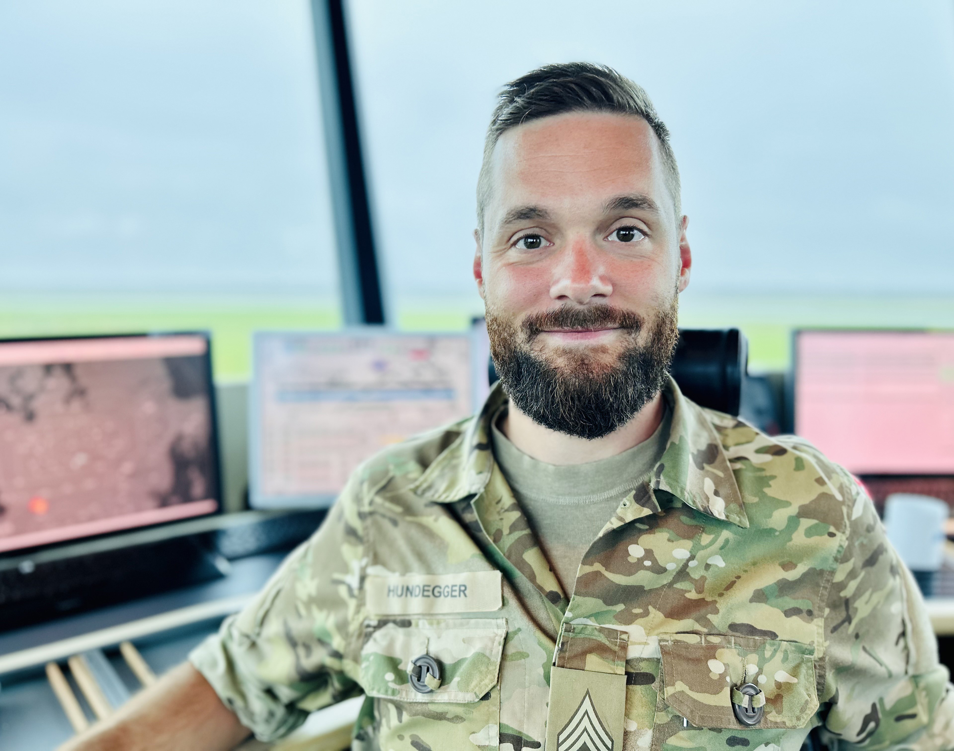 En uniformeret medarbejder sidder i et kontroltårn foran flere skærme og smiler mod kameraet.