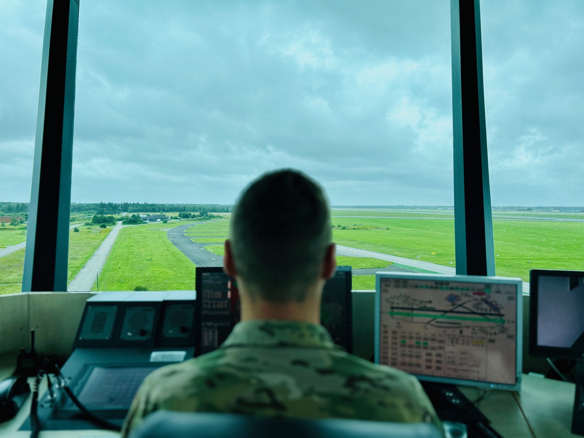 En flyveleder sidder i et kontroltårn og overvåger landingsbaner og grønne arealer gennem store vinduer, mens radarskærme og instrumenter er foran ham.
