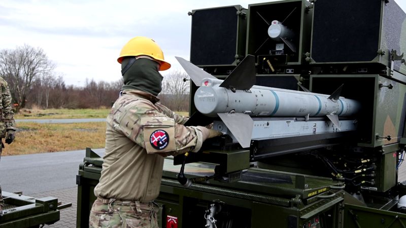 Soldat i camouflageuniform og gul hjelm håndterer et missil ved en affyringsrampe på et militært køretøj udendørs.