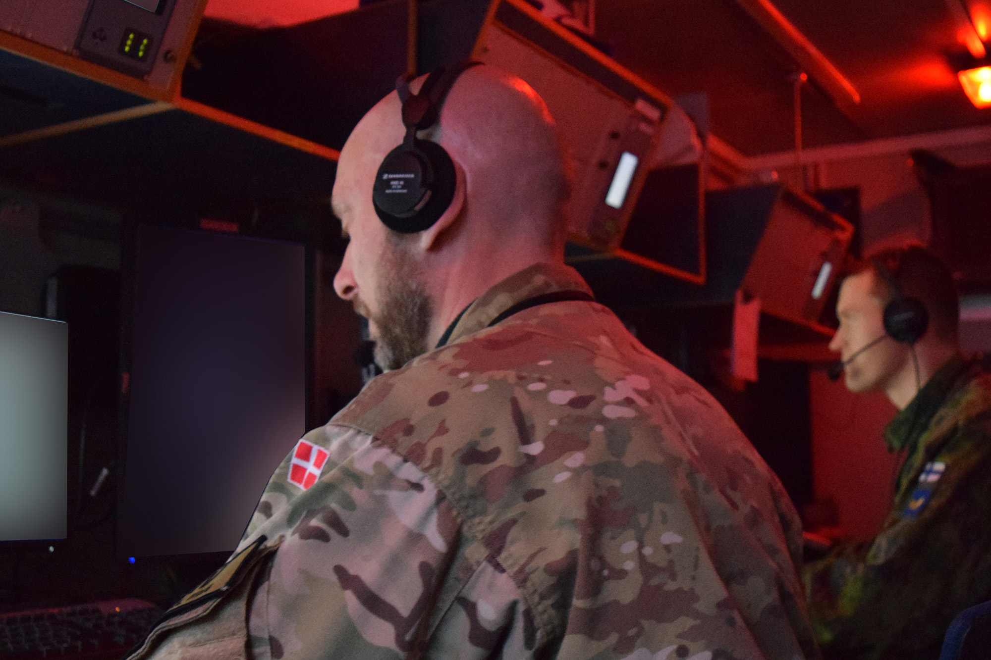 To soldater i uniform sidder ved computerskærme i en operationscentral med rødt lys og headset på.