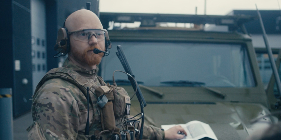 Soldat i camouflage med headset og beskyttelsesbriller står foran et militærkøretøj og ser til siden.