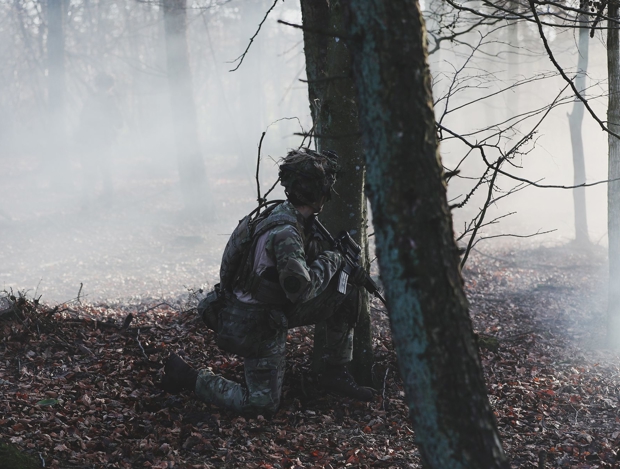 soldat i camouflage sider knælende bag træ i tåget skov med geværet i hånden