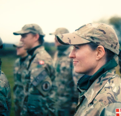 Soldater i camouflageuniform står på række under øvelse i felten. En kvindelig soldat i forgrunden smiler og ser fremad.