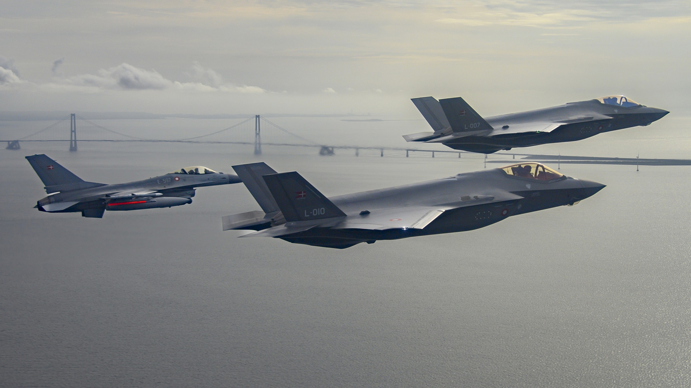 Tre danske kampfly flyver i formation over Storebæltsbroen på en overskyet dag.