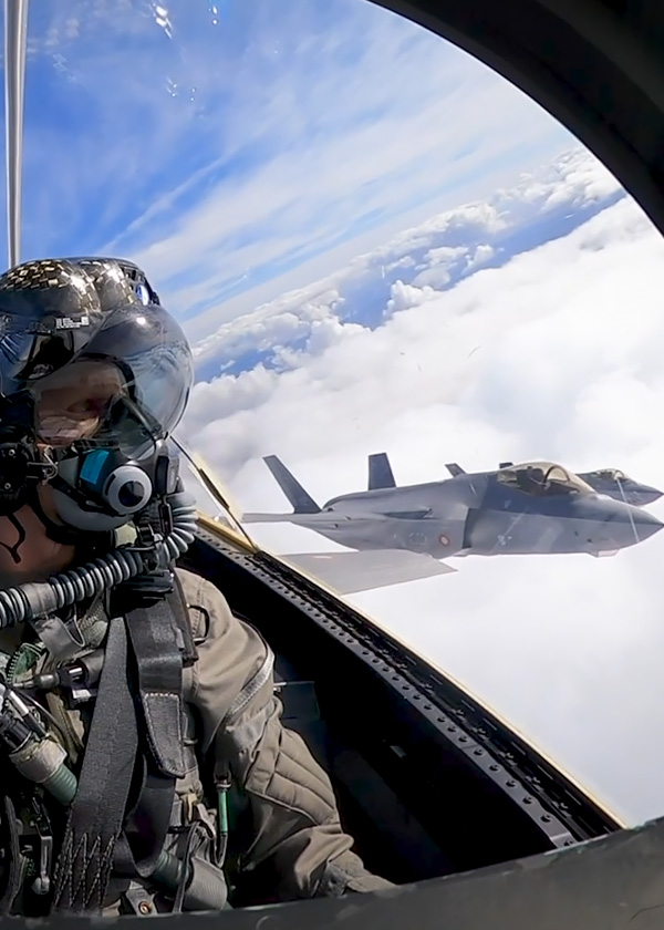 En jagerpilot i cockpittet flyver over skyerne med et dansk F-35 kampfly i baggrunden.