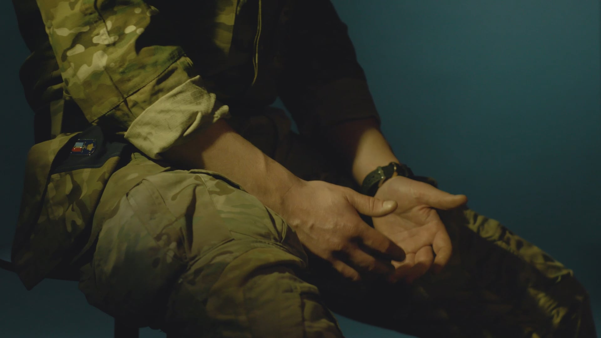 Person i militæruniform med foldede hænder i skødet, set i dæmpet lys.