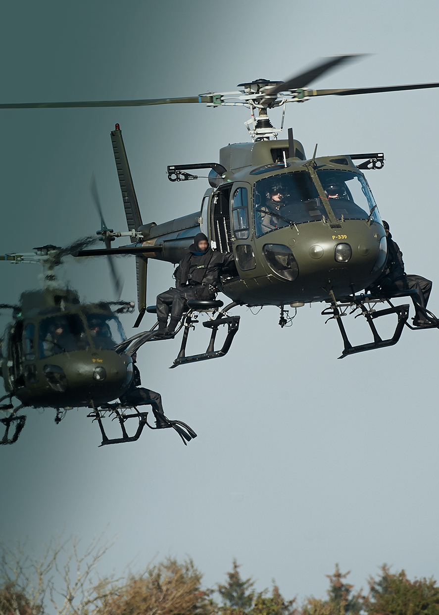 To militærhelikoptere i luften med specialstyrker siddende uden på trinbrættene, klar til indsættelse