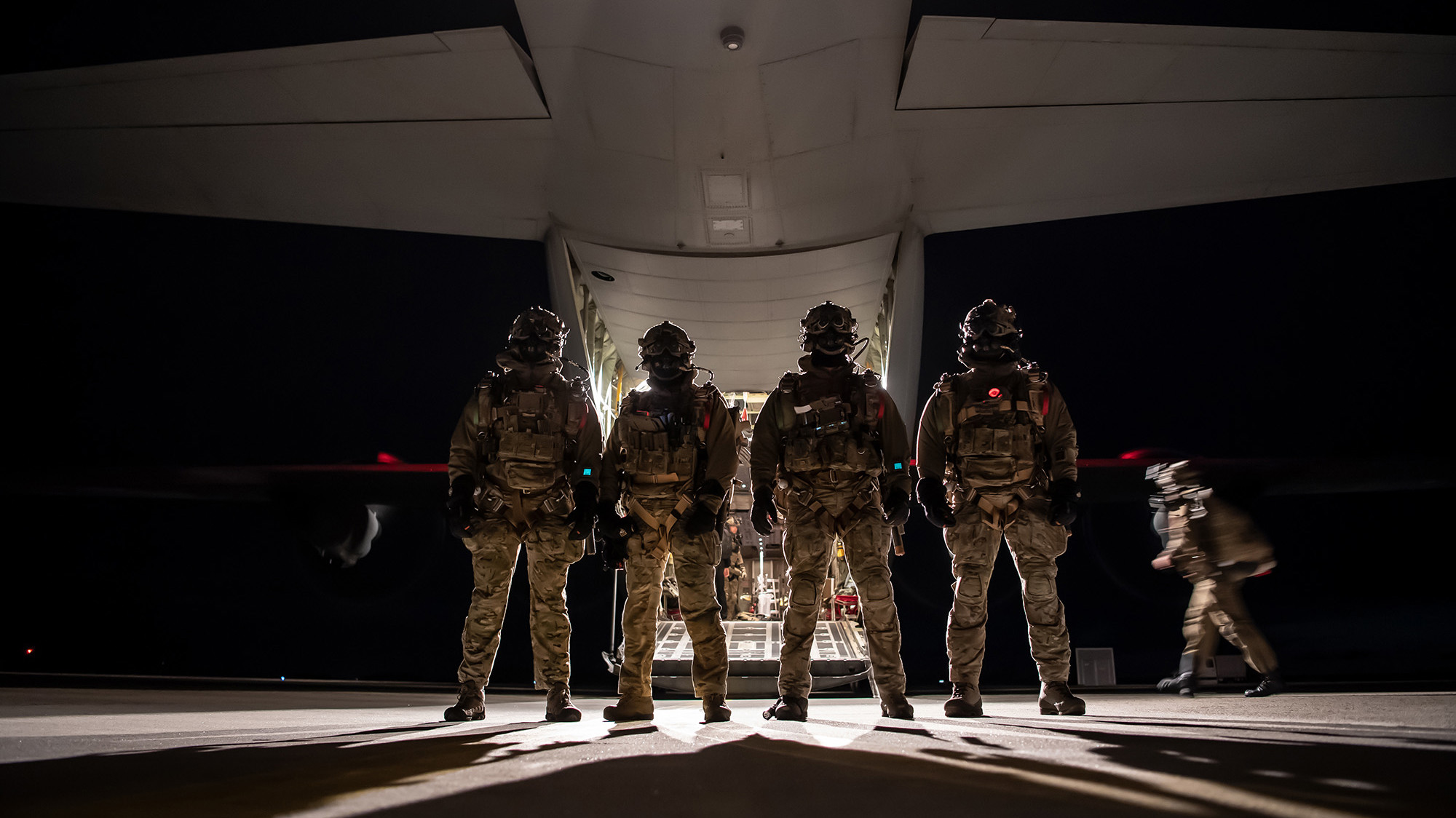 Fem soldater i fuld kampuniform står opstillet foran et militærflys åbne lastrampe om natten, klar til indsættelse.