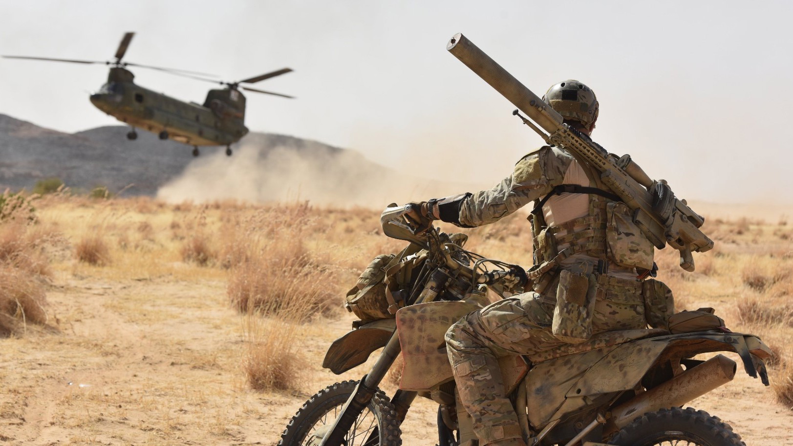 Soldat på terrængående motorcykel i ørkenlandskab observerer en lavtflyvende Chinook-helikopter.