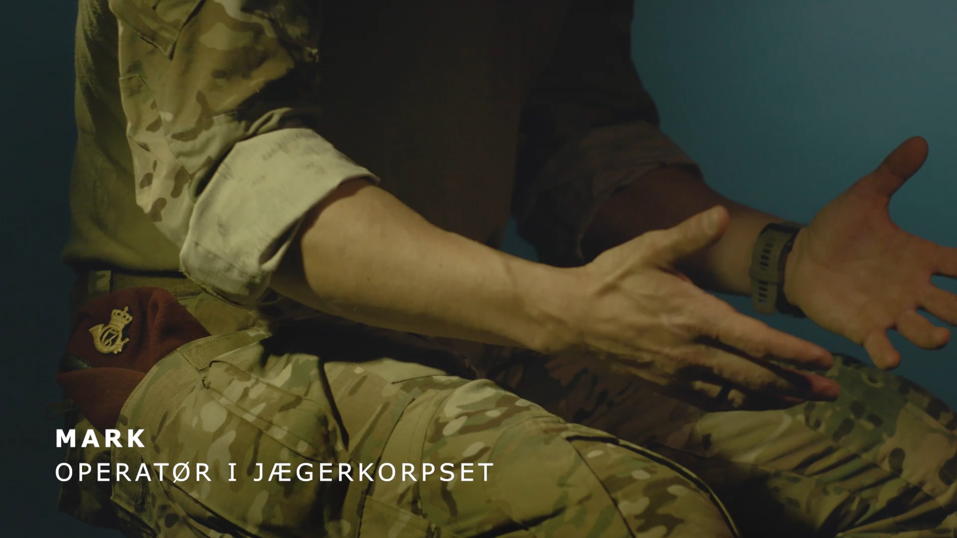 Person i camouflageuniform med bordeaux baret og hænder i skødet.