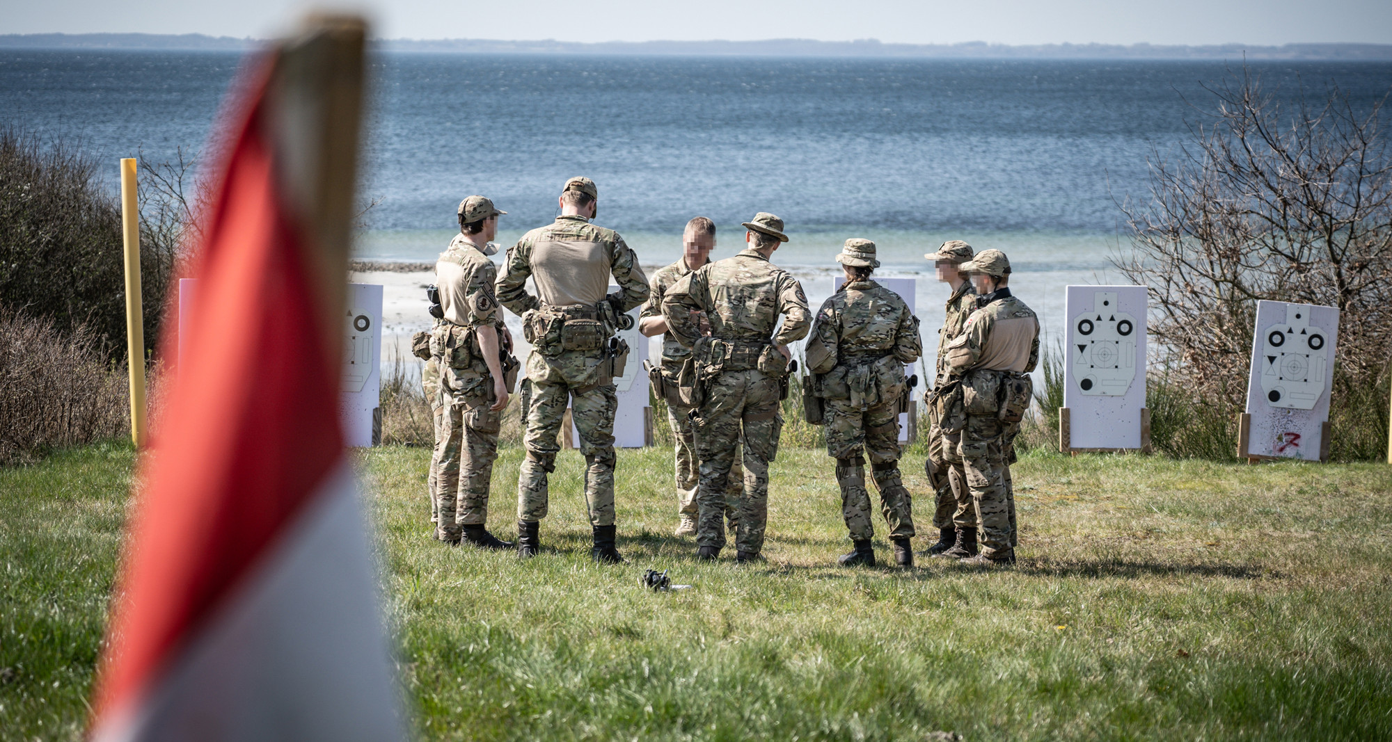 Danske soldater i uniform til skydeøvelse ved kysten, samlet foran målskiver med havet i baggrunden.