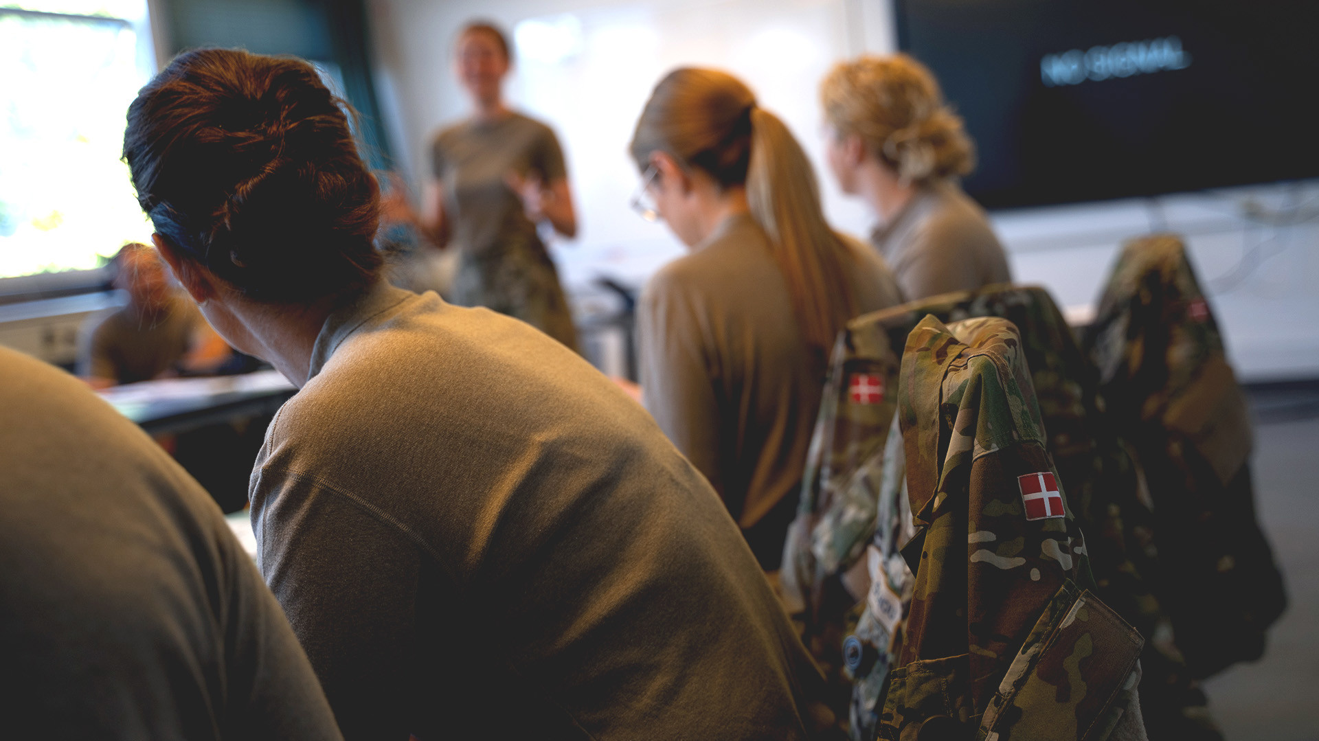 Undervisningssituation, hvor soldater i uniform følger med, mens en instruktør taler foran klassen.