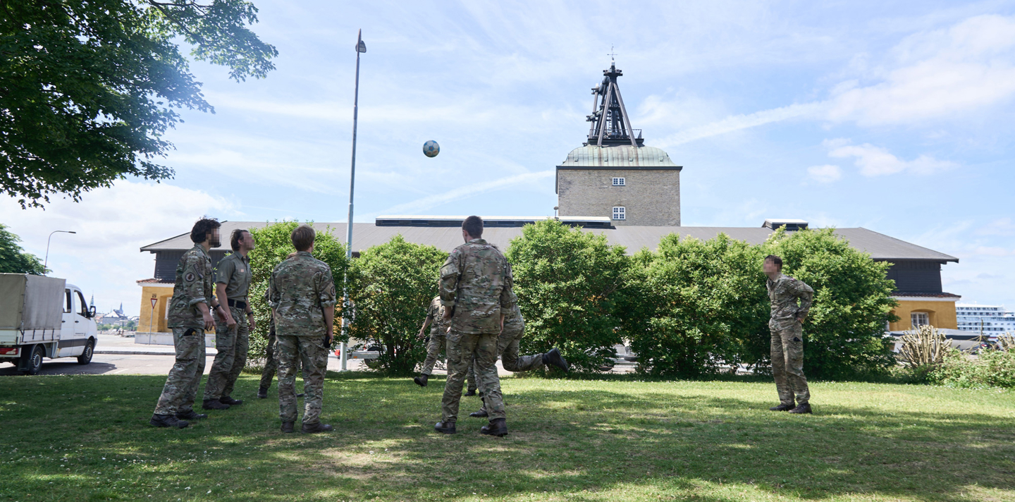 Soldater i uniform spiller bold på en græsplæne foran en kasernebygning.
