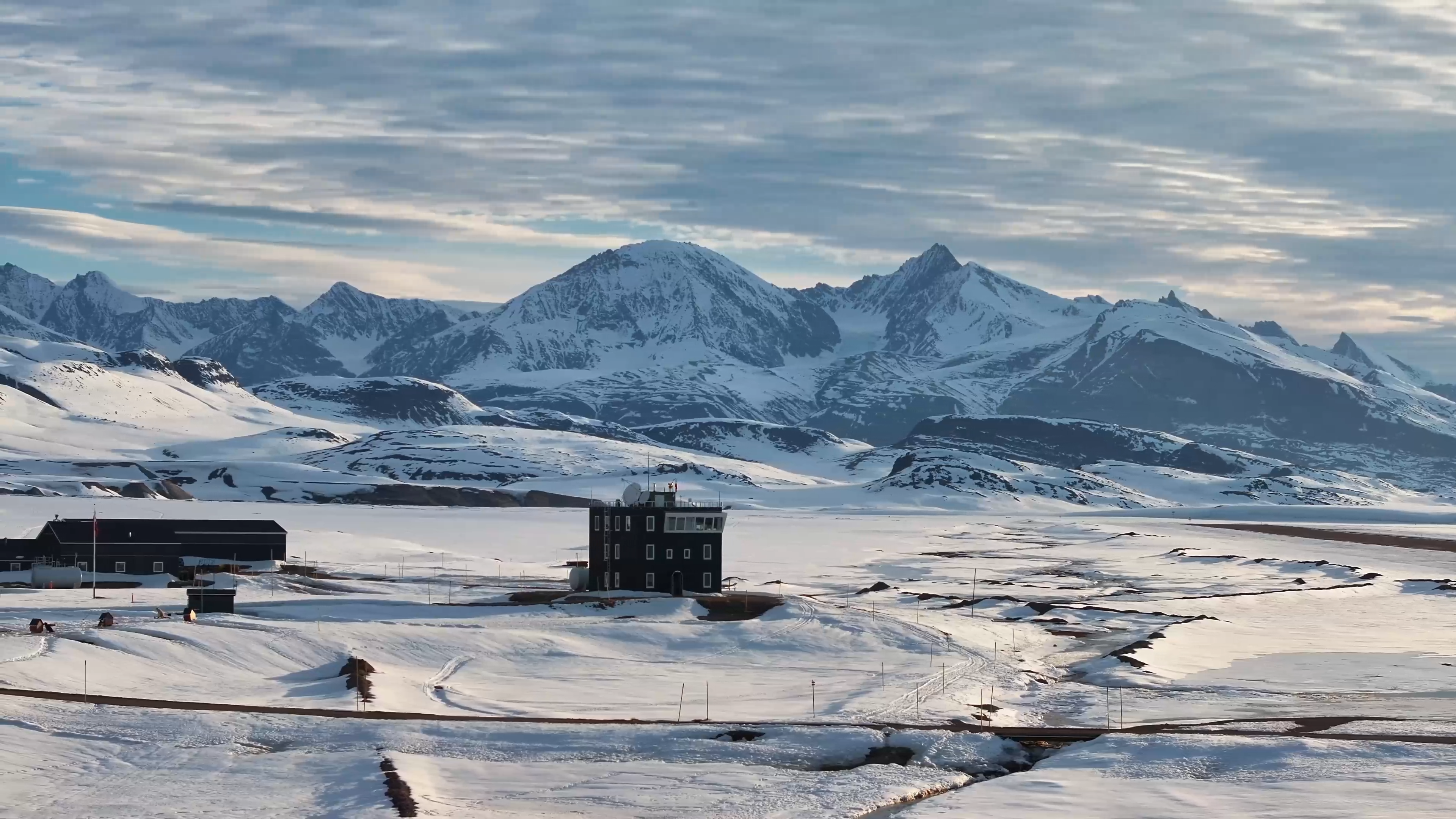 Sneklædte bjerge i Nordgrønland rejser sig dramatisk i baggrunden af et arktisk landskab med radarbygning og isdækkede vidder i forgrunden.