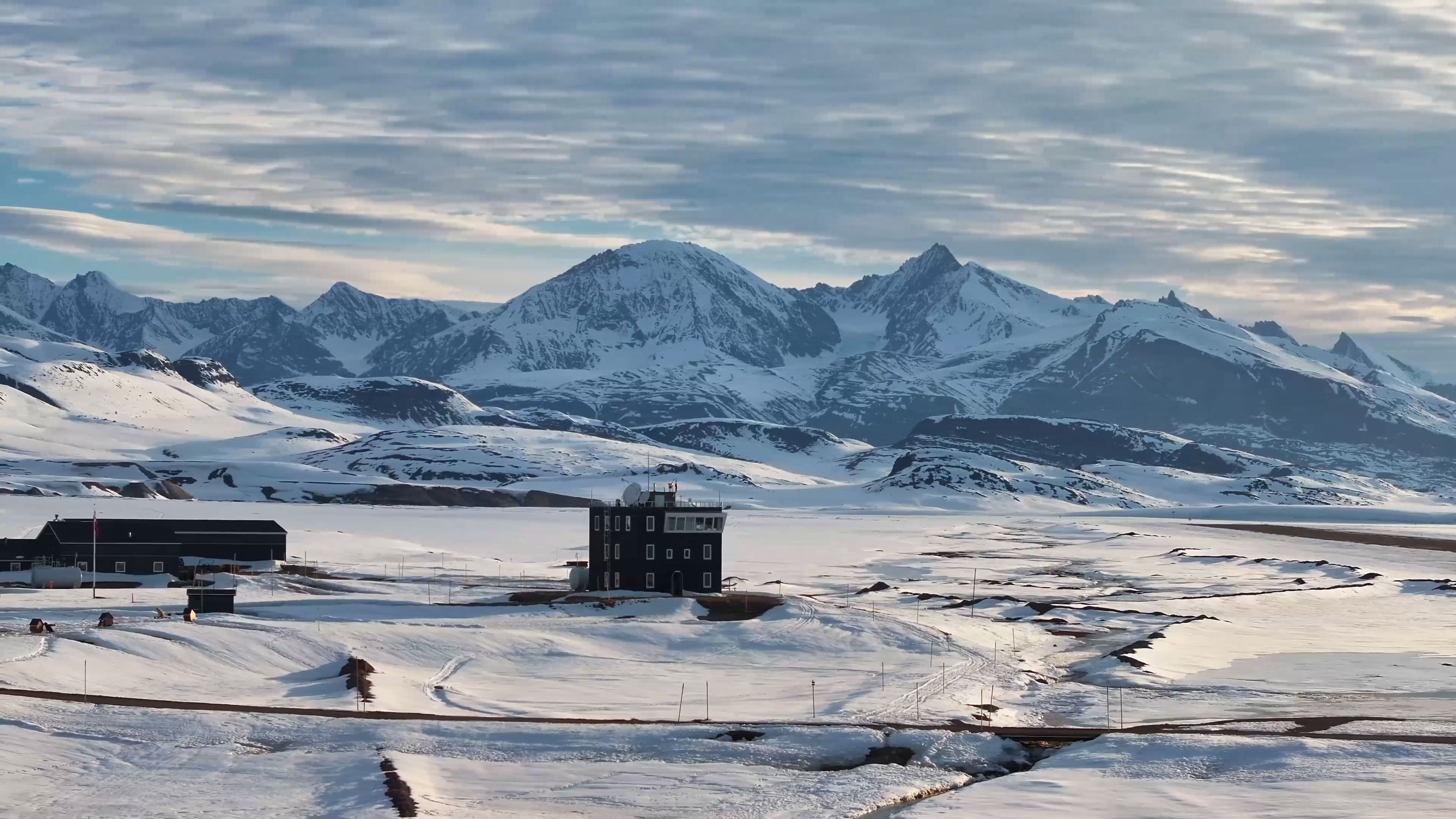 Sneklædte bjerge i Nordgrønland rejser sig dramatisk i baggrunden af et arktisk landskab med radarbygning og isdækkede vidder i forgrunden.