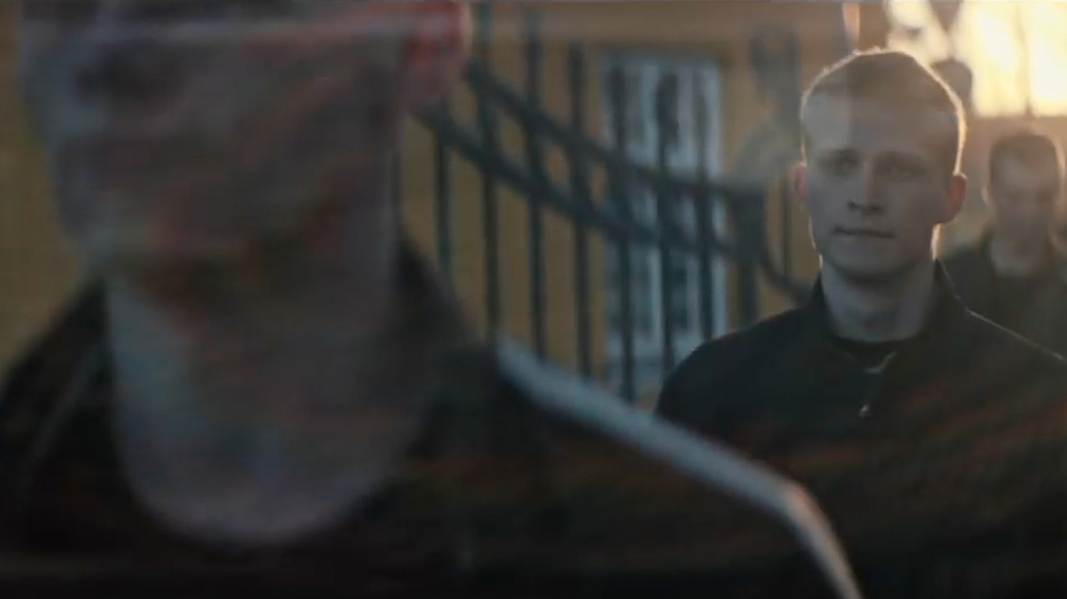 En gruppe personer går udendørs i modlys, hvor den forreste er i fokus, mens andre bevæger sig bagved langs et hegn.