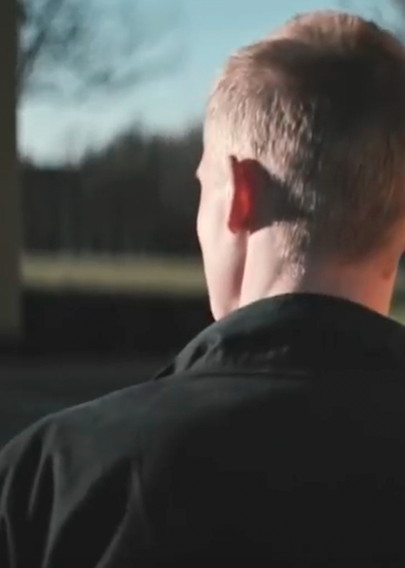 En person set bagfra står udendørs i sollys, med kort hår og mørk jakke, mens baggrunden er let sløret.