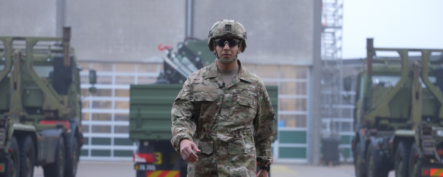 Hav går på en kaserne med uniform på, hvor der bag ham er militære køretøjer