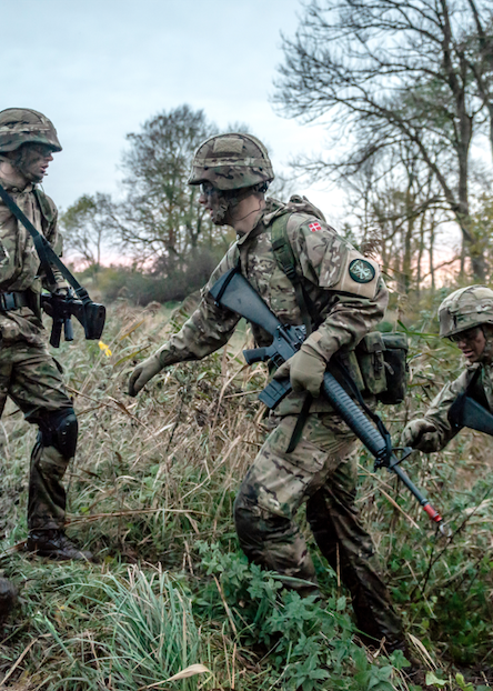 Soldater i camouflageuniform bevæger sig gennem højt græs og ujævnt terræn under en øvelse i naturen.
