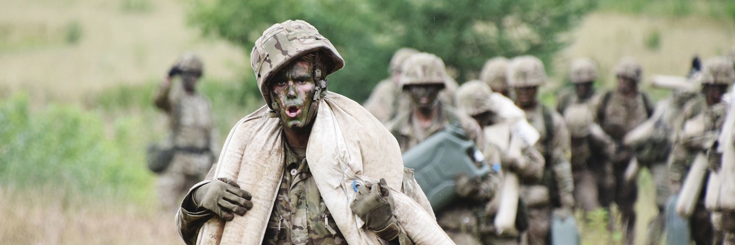 En soldat bærer sandsække under en øvelse, mens en enhed følger efter i baggrunden.