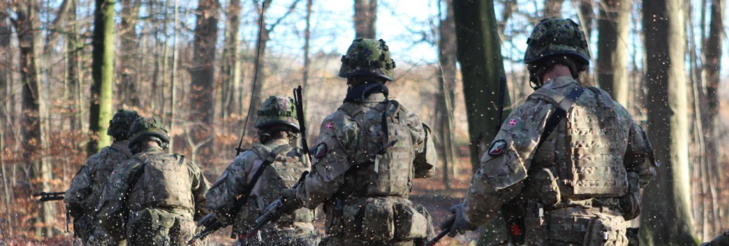 En gruppe soldater i uniform løber gennem lavt vand i en skov, så vandet sprøjter omkring dem, mens de bevæger sig fremad med deres udrustning og våben.