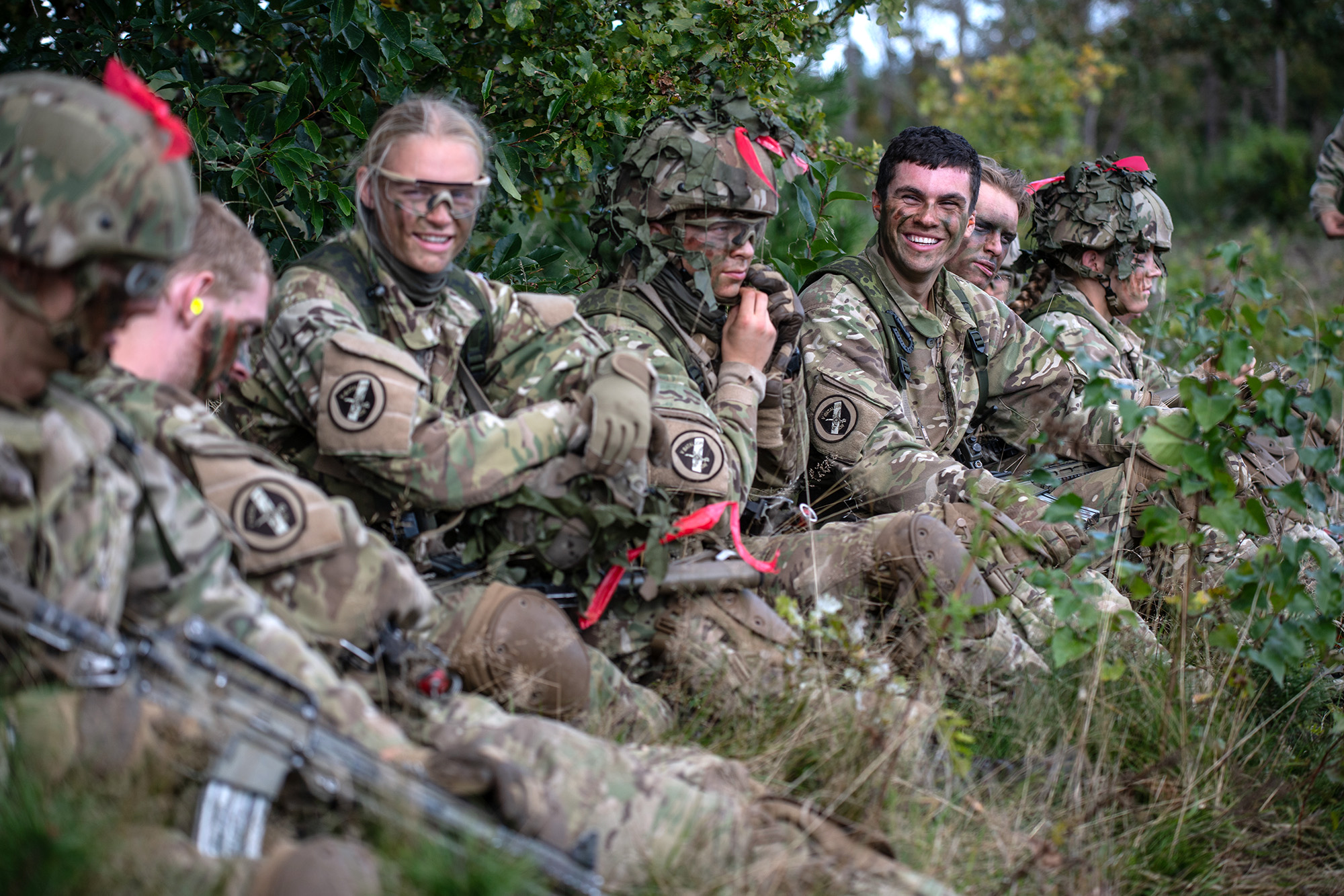 Værnepligtige i camouflageuniformer sidder i terrænet og holder pause med smil og kammeratlig stemning.